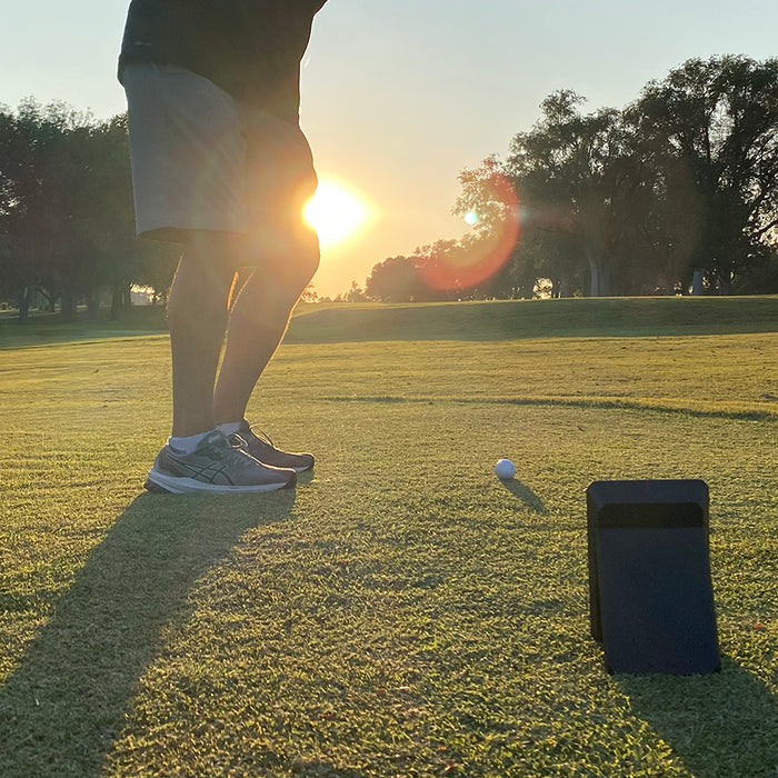 The lower half of golf reviewer Marc with a sunset at the golf range in the background and a Swing Caddie SC4 in the foreground as he gets ready to swing