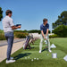 two men at a driving range standing outside about to swing a golf club and hit a golf ball with skytrak+ golf simulator and golf launch monitor on the ground next to them on a golf mat