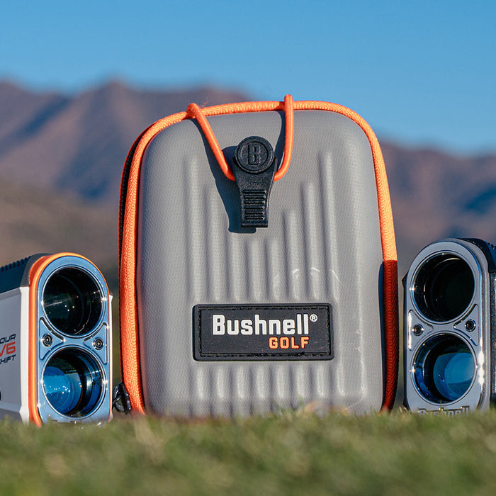 Two Bushnell Tour V6 golf rangefinders with the carrying case between them on the grass outdoors with a mountain range in the background