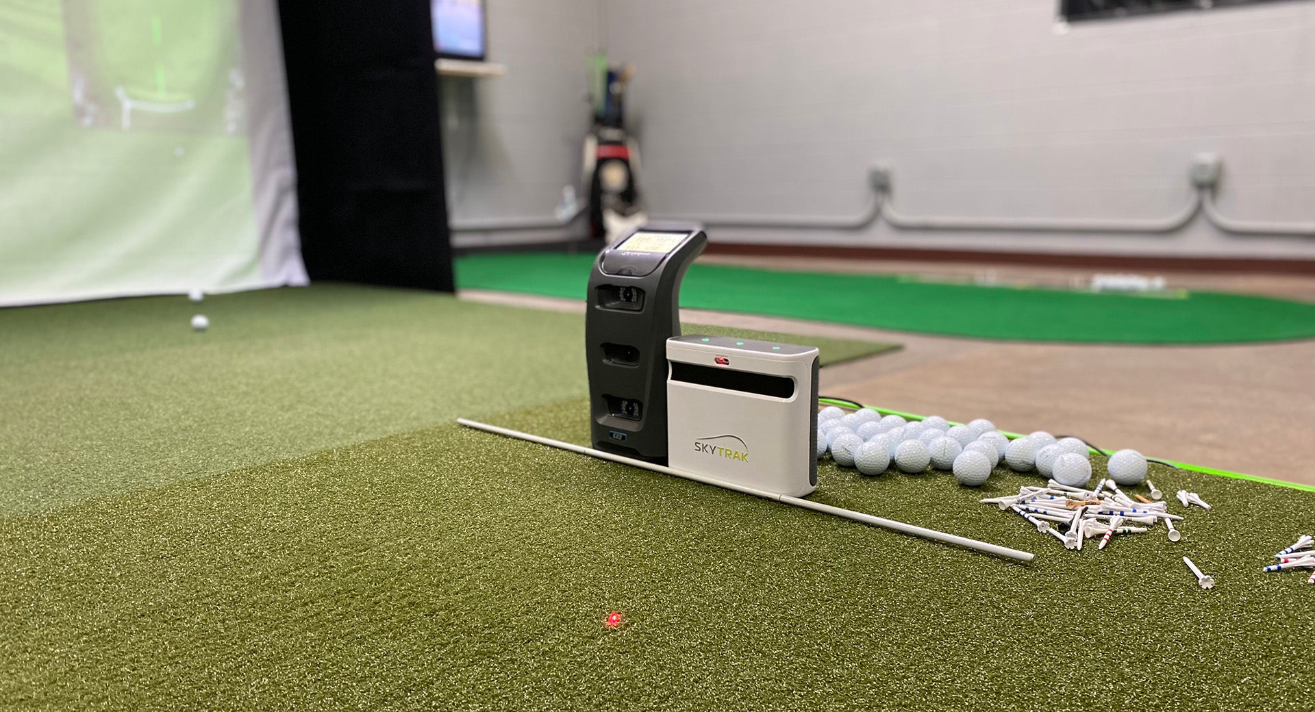 The SkyTrak+ launch monitor next to the Foresight Sports GC3 next to many golf balls and golf tees on a golf mat in an indoor simulator