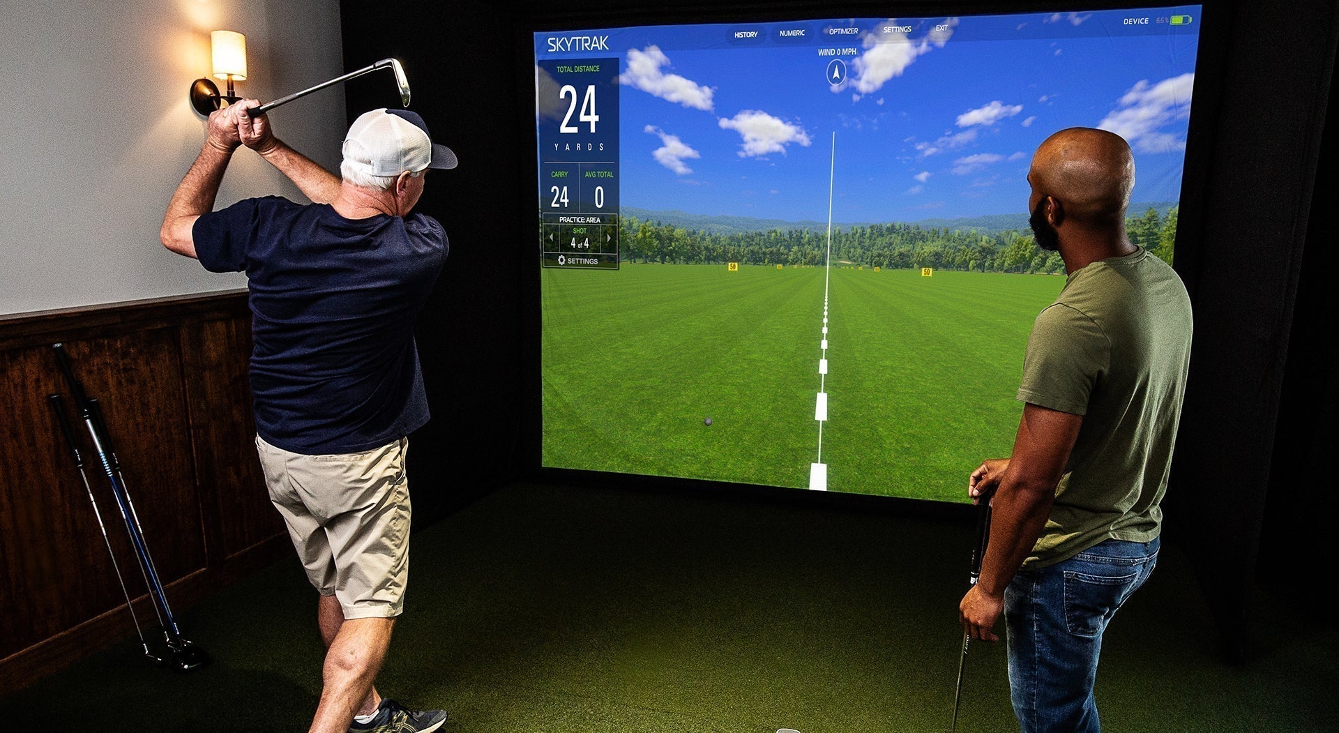 Two men, one swinging a golf club, in a home golf simulator with SkyTrak software on the impact screen in front of them