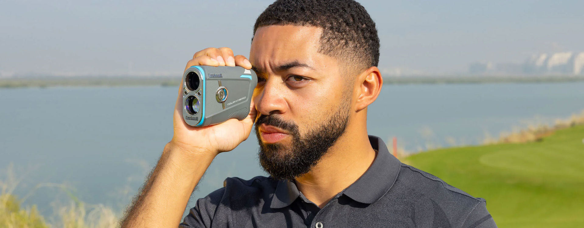 A golfer with a Bushnell Tour Hybrid rangefinder to his eye at a golf course near a body of water