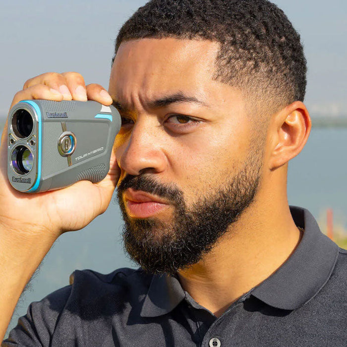 A golfer with a Bushnell Tour Hybrid rangefinder to his eye at a golf course near a body of water