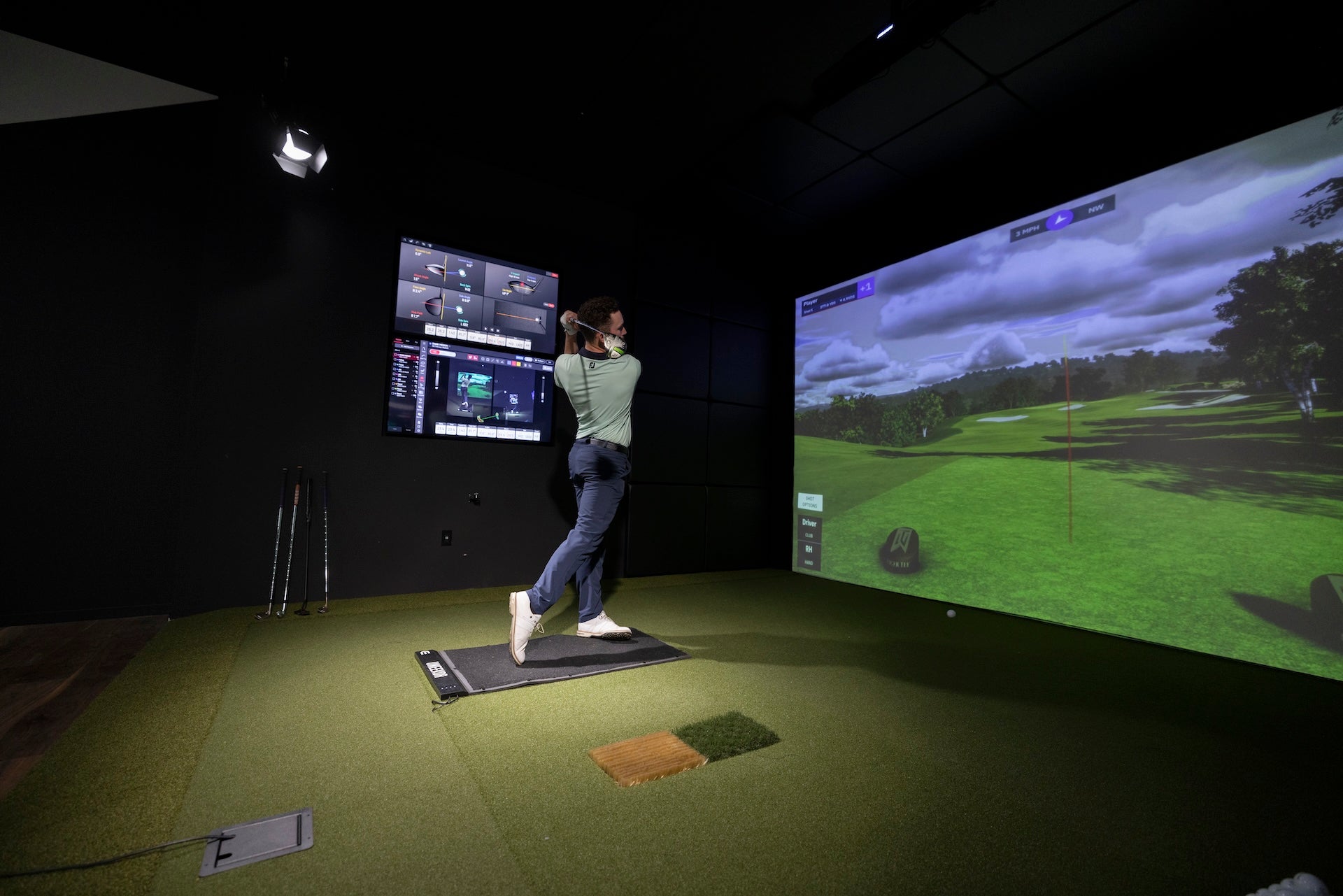 A golfer in a Uneekor golf simulator with virtual golf on the impact screen and data showing on flatscreens off to the side