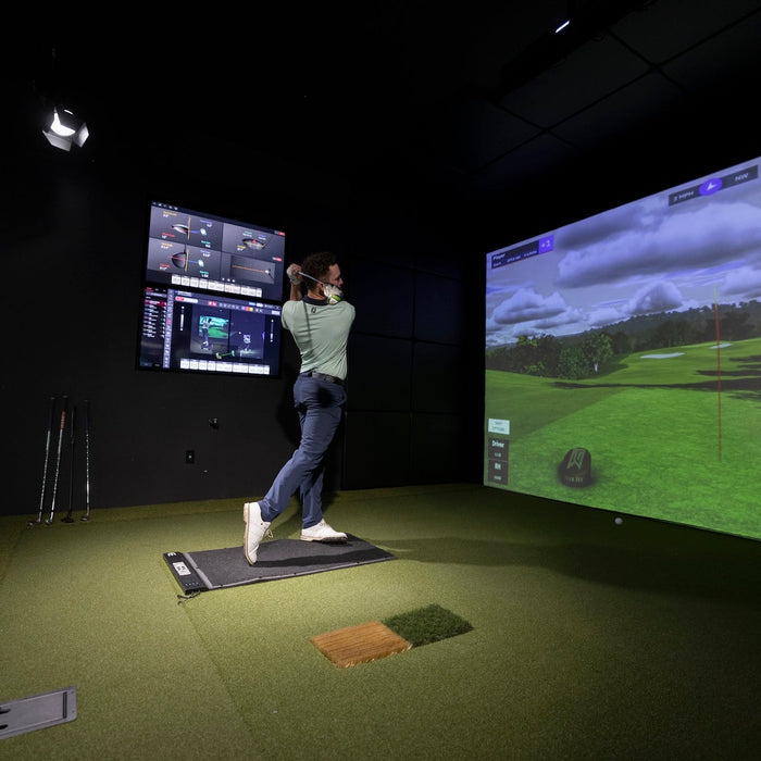 A golfer in a Uneekor golf simulator with virtual golf on the impact screen and data showing on flatscreens off to the side