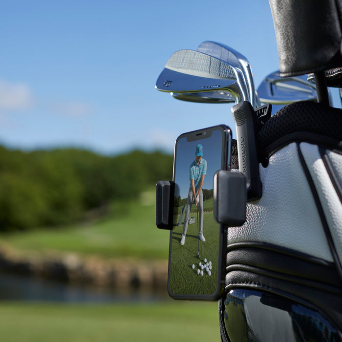 The Garmin Golf app on a smartphone clipped to a golf bag showing video of a golfer from front view getting ready to swing