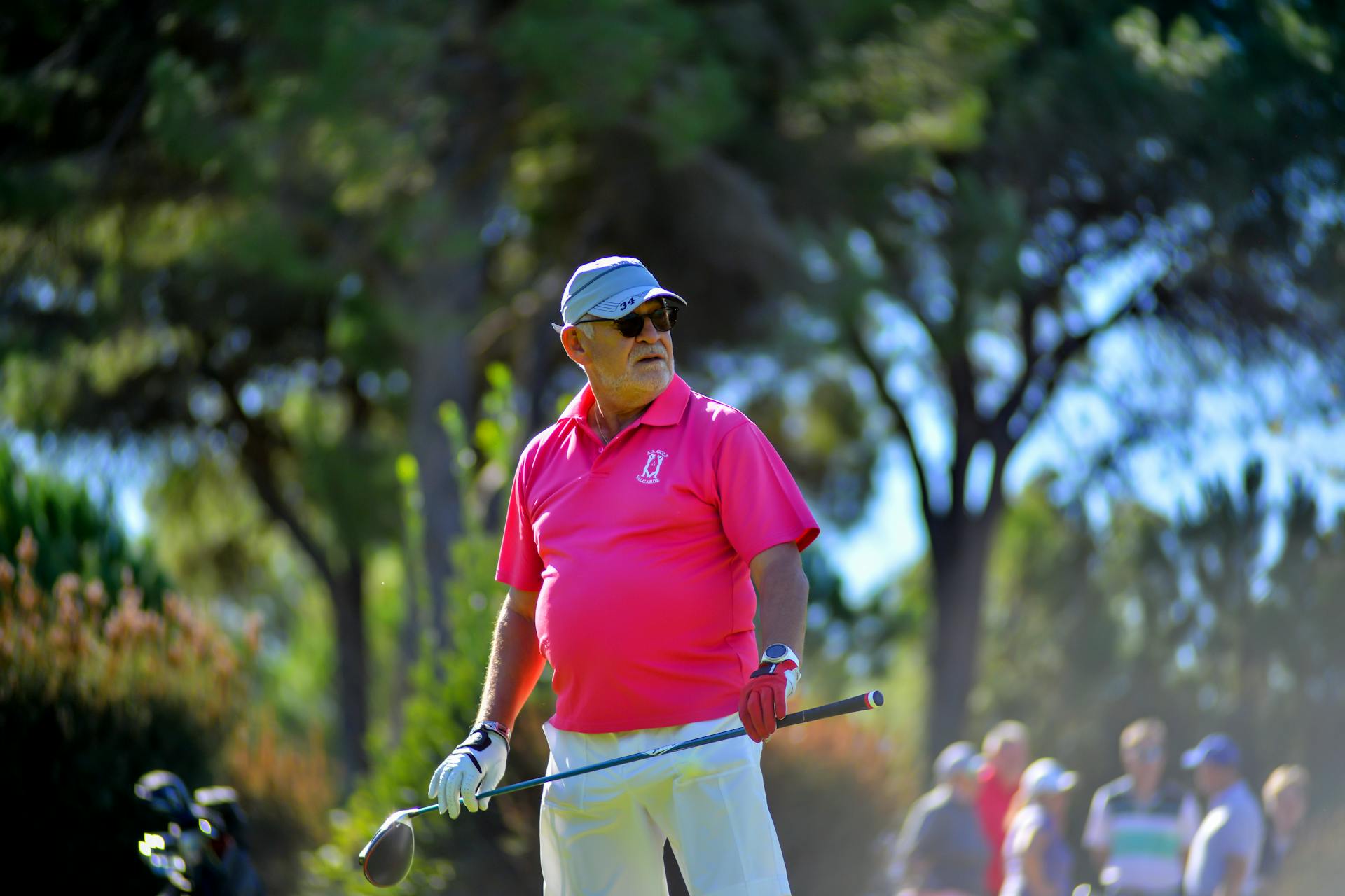 Senior golfer in a pink shirt and white hat and pants holding a driver with both hands and looking out at the course from the tee