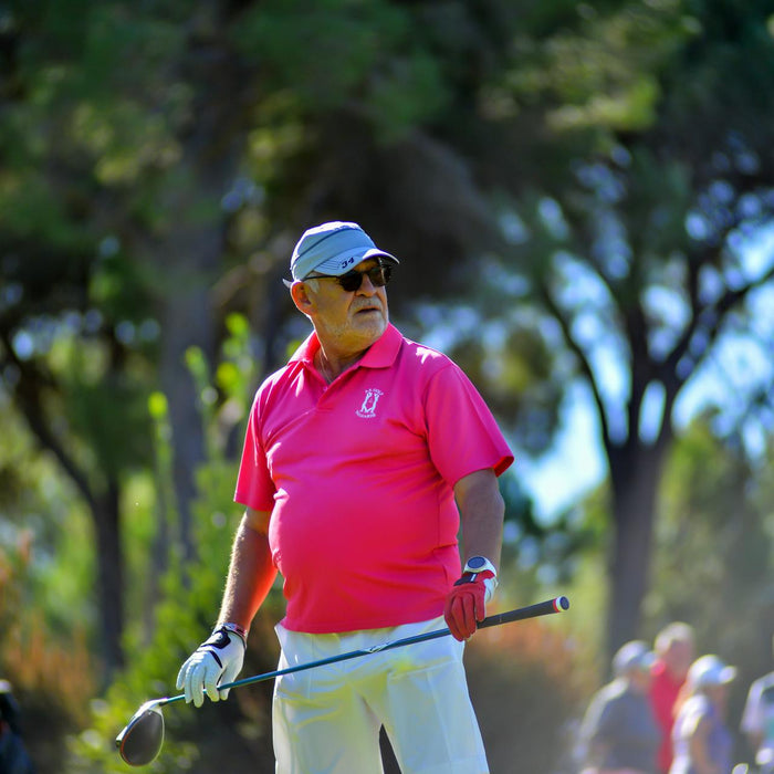 Senior golfer in a pink shirt and white hat and pants holding a driver with both hands and looking out at the course from the tee