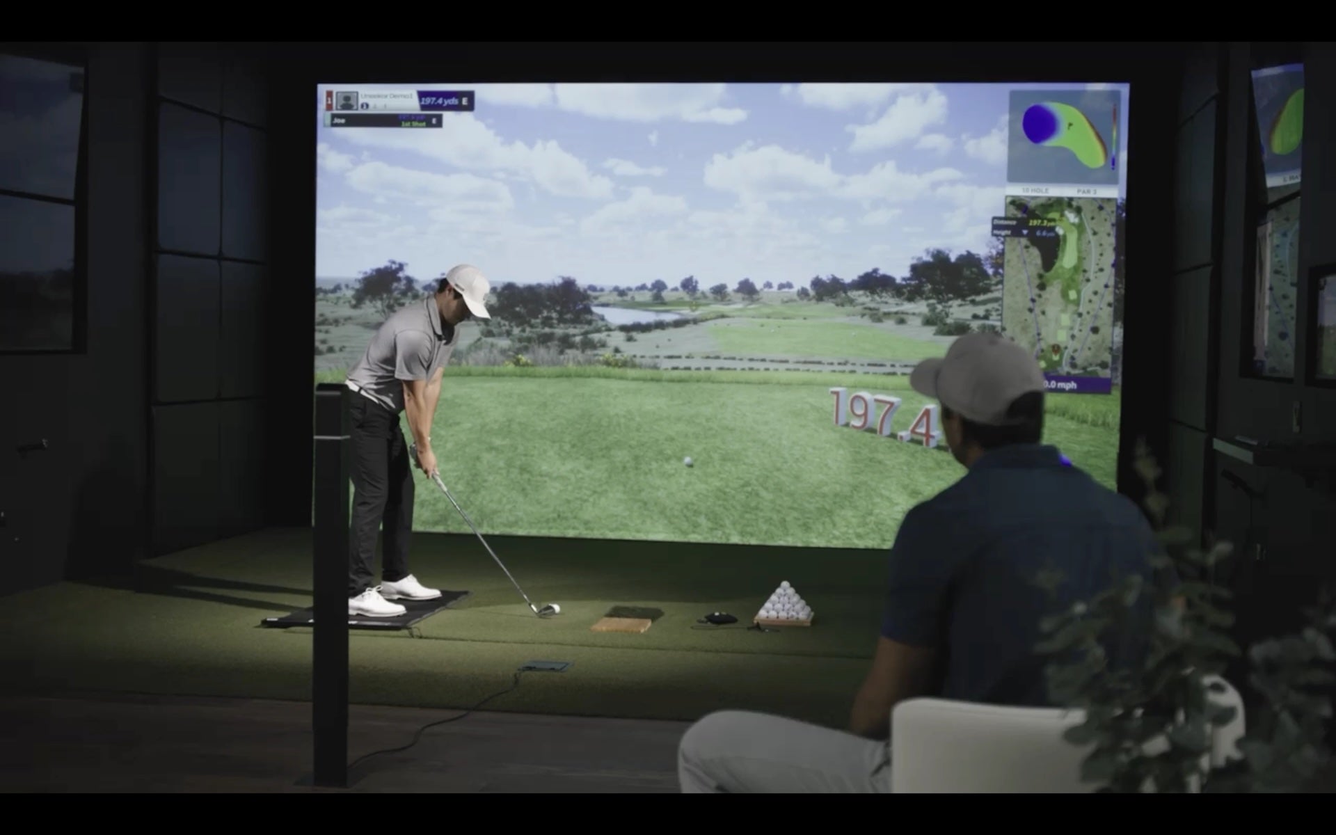 A Uneekor golf simulator with a golfer preparing to swing and a man behind him sitting in a chair and watching