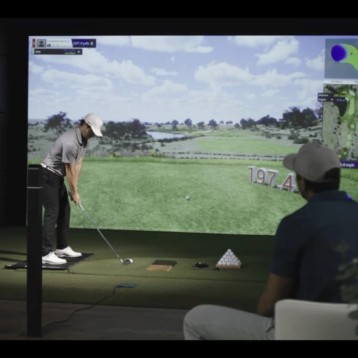 A Uneekor golf simulator with a golfer preparing to swing and a man behind him sitting in a chair and watching