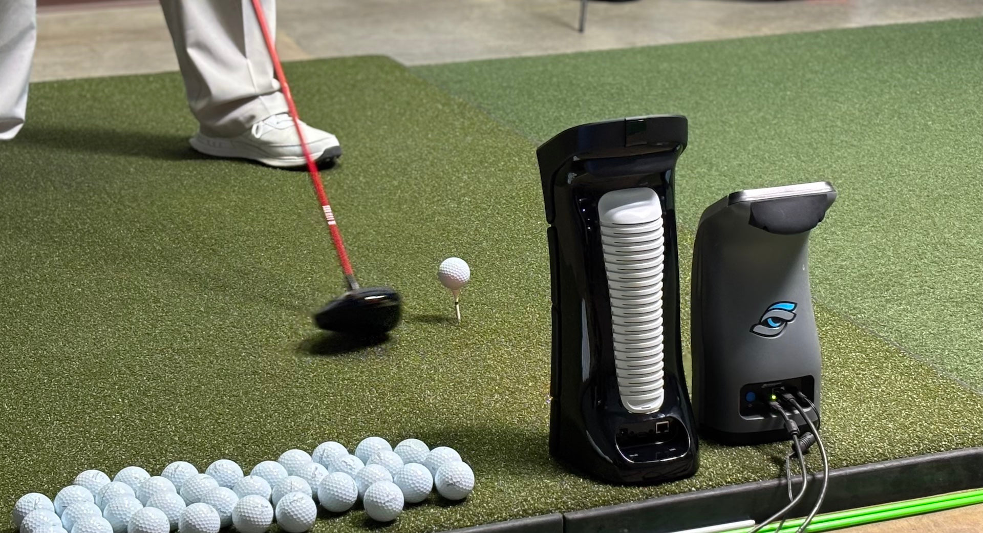 The backs of the Uneekor Eye Mini and Foresight GC3 next to golf balls and a driver and ball on a tee on a golf hitting mat in a golf simulator