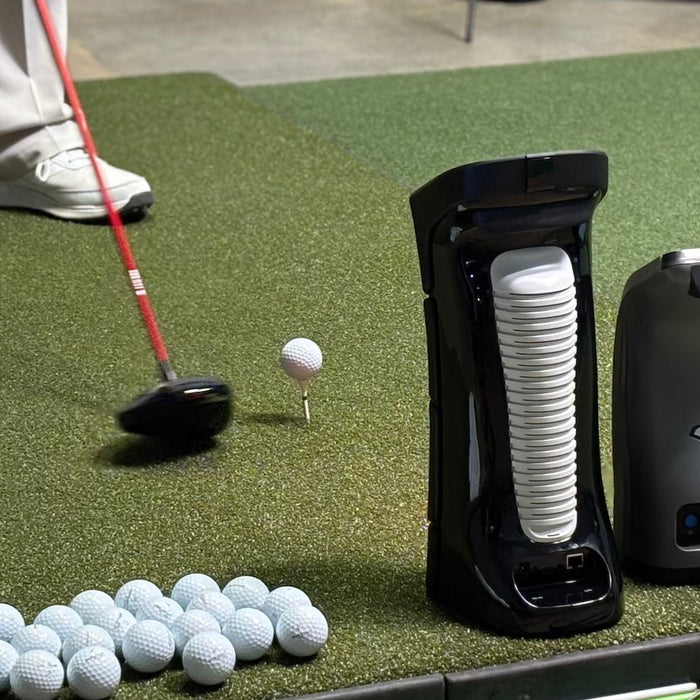 The backs of the Uneekor Eye Mini and Foresight GC3 next to golf balls and a driver and ball on a tee on a golf hitting mat in a golf simulator