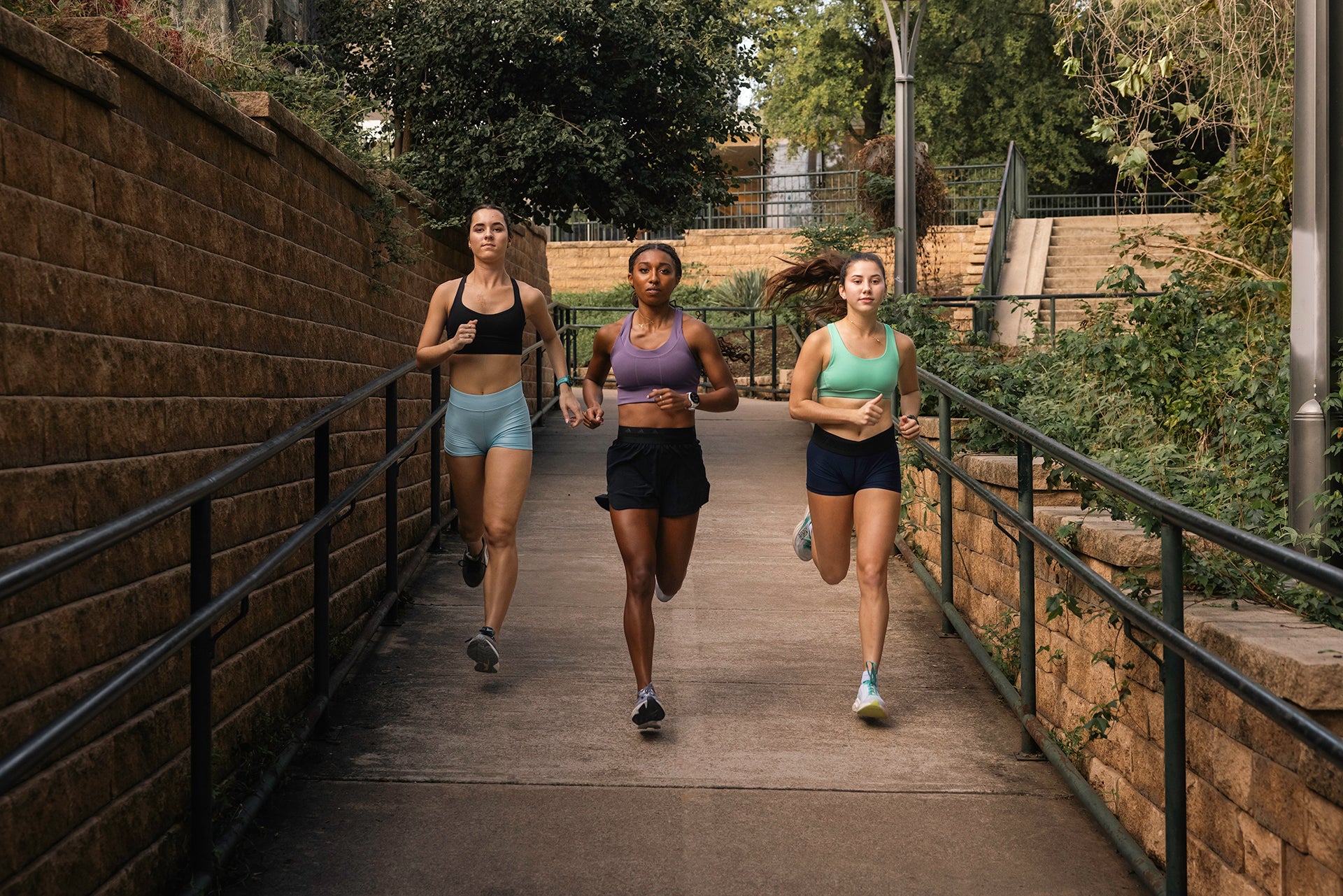 Three women running down a sidewalk next a rock wall and railings with Forerunner 165 running watches on