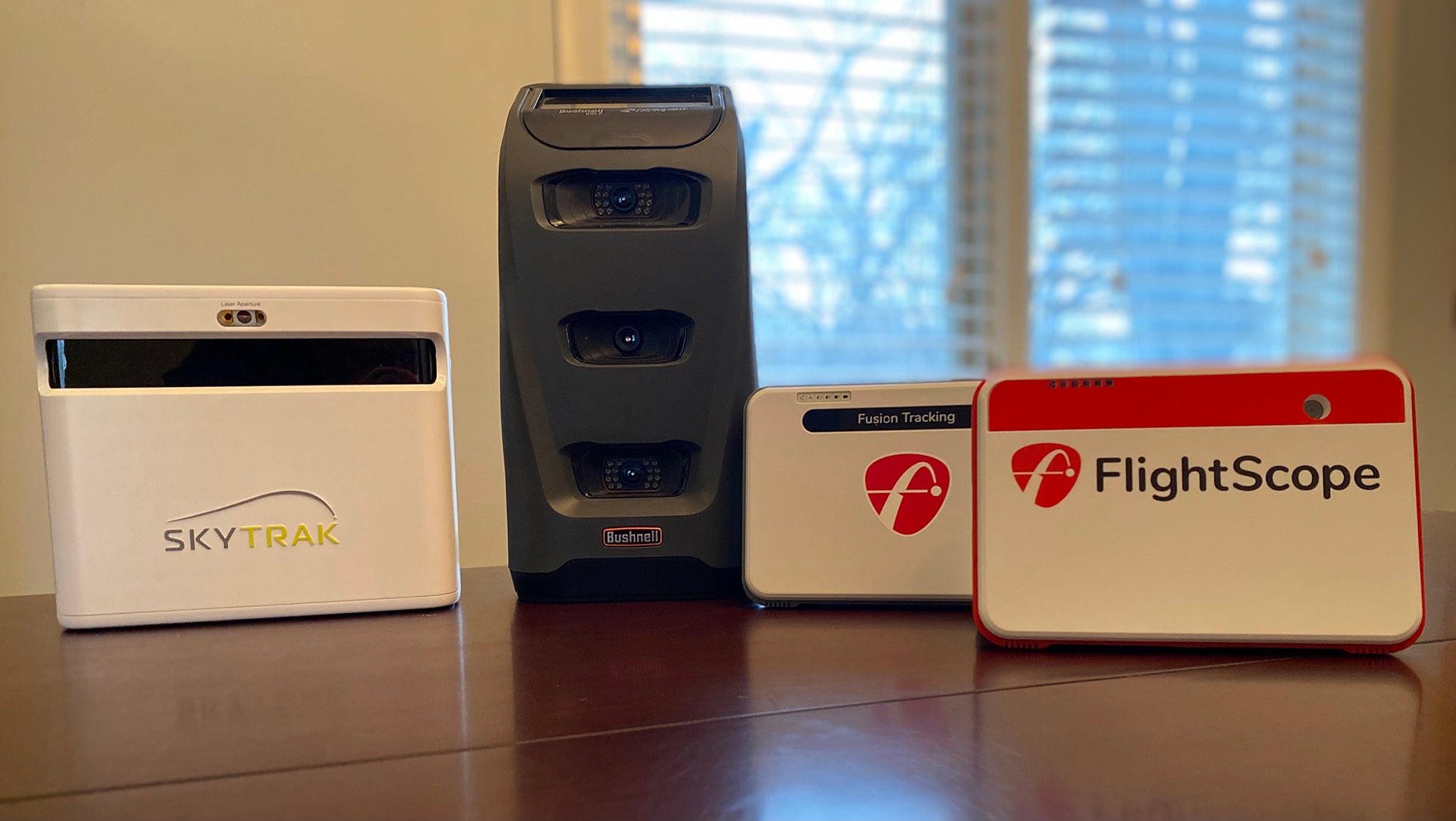 The SkyTrak+, Bushnell Launch Pro, FlightScope Mevo+ Limited Edition, and Mevo+ launch monitors standing together on a table with a window in the background