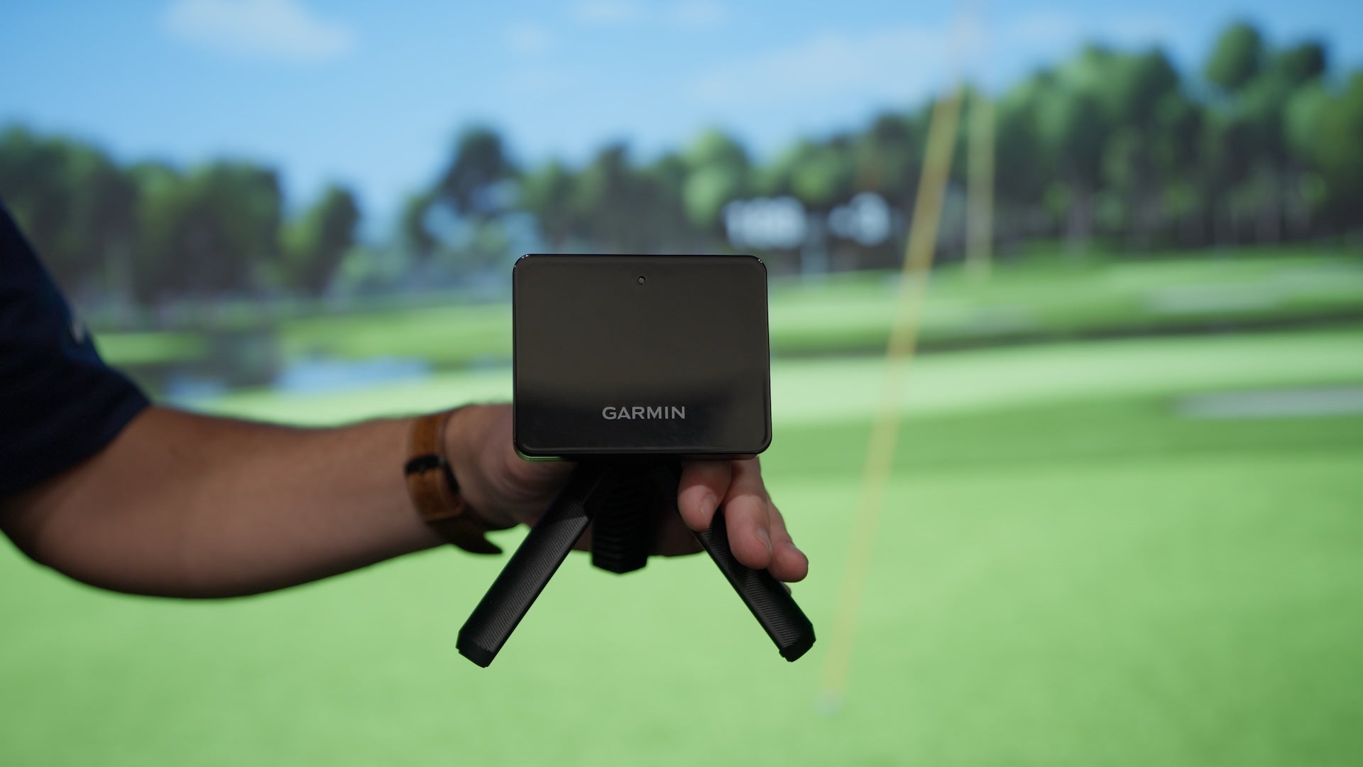 Garmin R10 in a hand in held in front of a golf impact screen with Home Tee Hero virtual golf on it