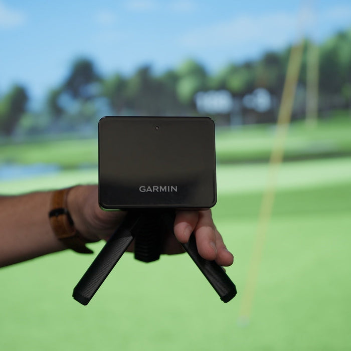 Garmin R10 in a hand in held in front of a golf impact screen with Home Tee Hero virtual golf on it