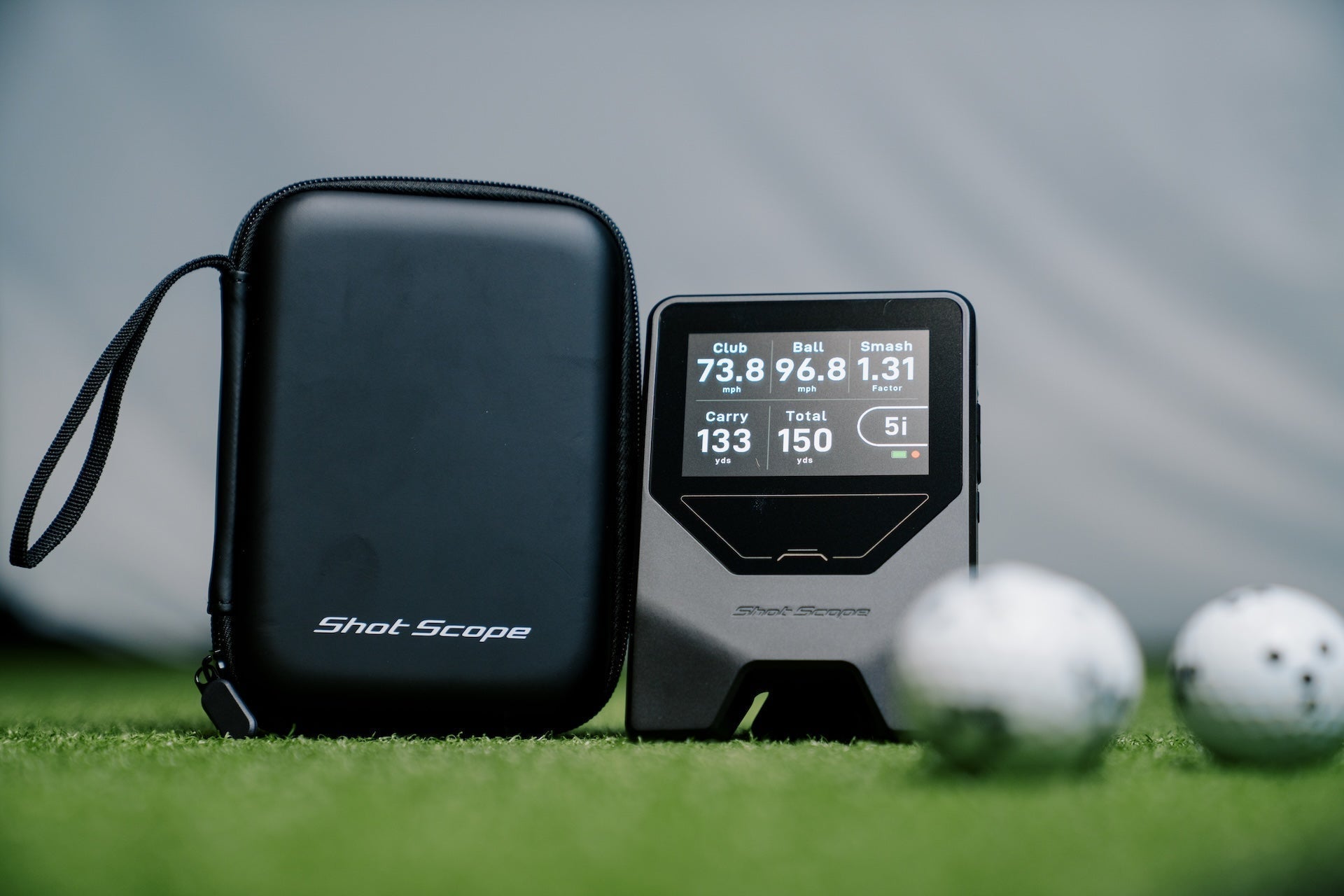 The Shot Scope LM1 on a golf hitting mat next to its carrying case and some golf balls