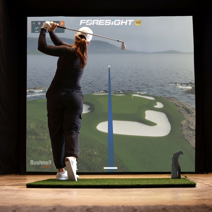 A woman golfer post swing in a golf simulator with the Bushnell Launch Pro and Foresight golf course on the impact screen