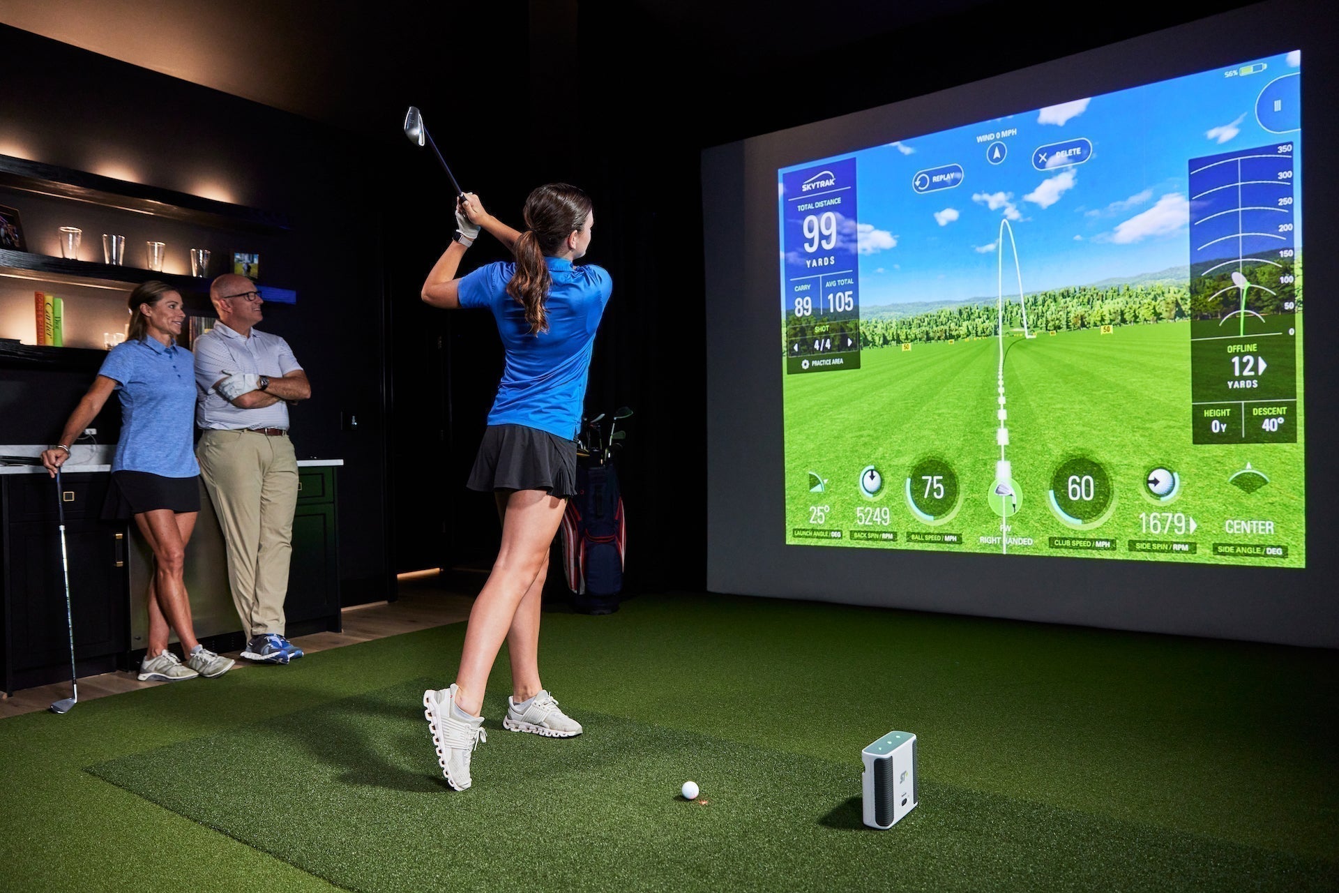 A girl post swing in a golf launch monitor with virtual golf on the impact screen while her parents look on from the side while leaning against a counter