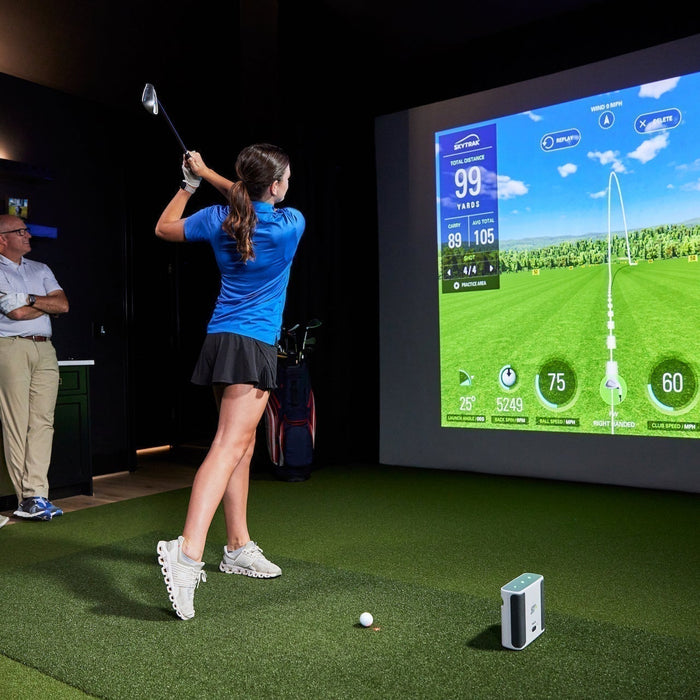 A girl post swing in a golf launch monitor with virtual golf on the impact screen while her parents look on from the side while leaning against a counter