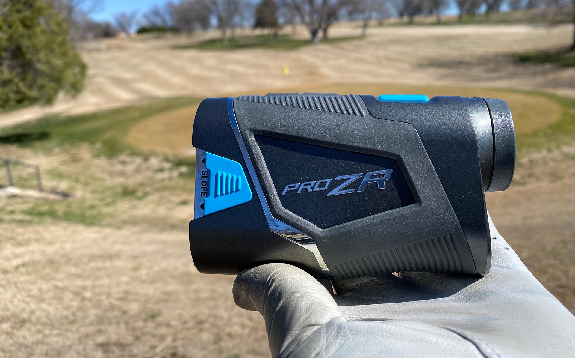 The Shot Scope PRO ZR golf rangefinder in a golf-gloved hand held up against a golf course in the background