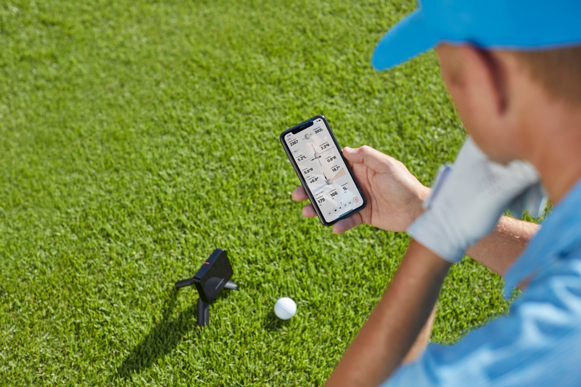 Golfer holding a smartphone with shot data on the display over a Garmin Approach R10 golf launch monitor and golf ball on a golf hitting mat