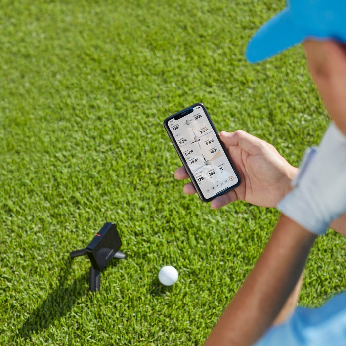 Golfer holding a smartphone with shot data on the display over a Garmin Approach R10 golf launch monitor and golf ball on a golf hitting mat