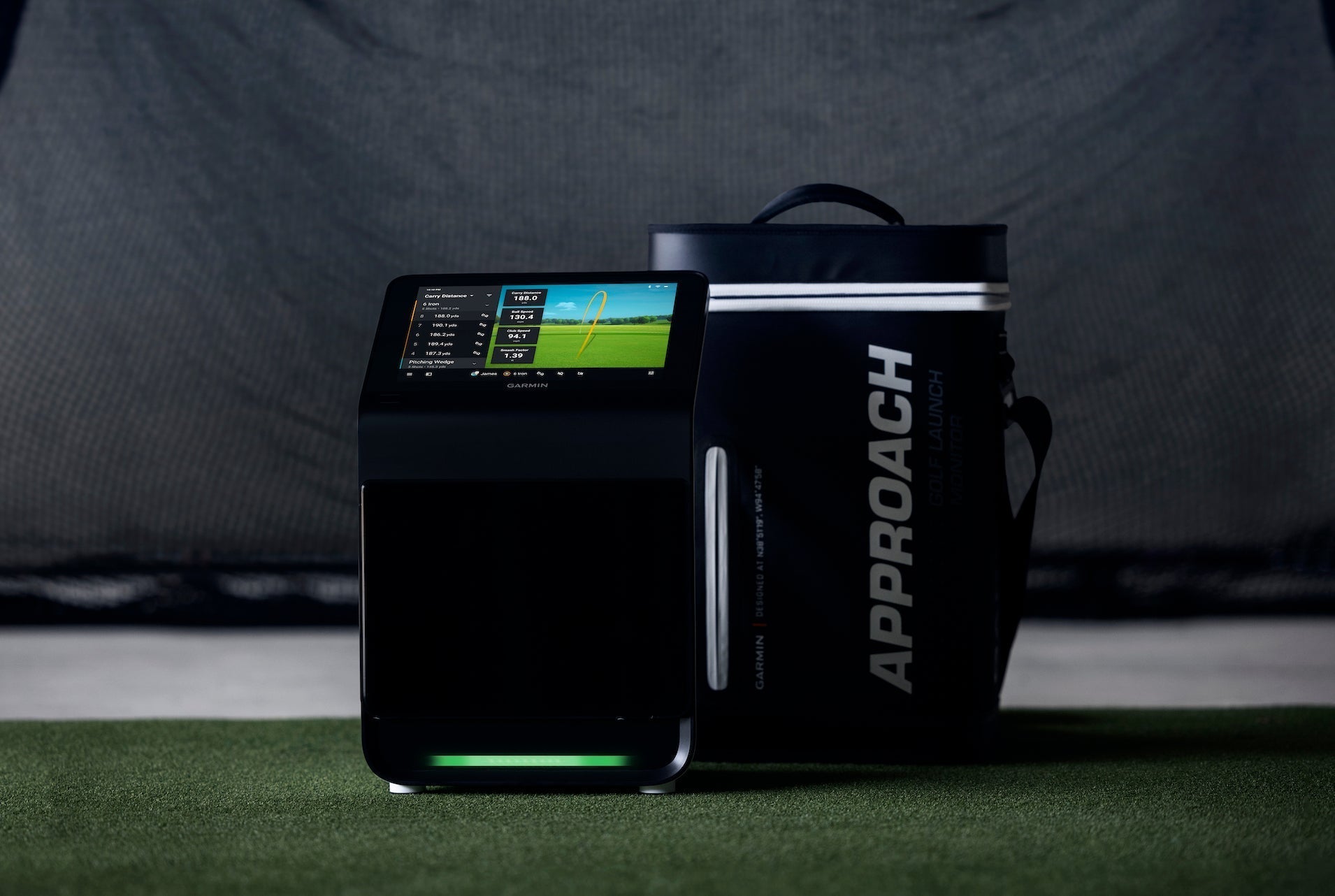 The Garmin Approach R50 next to its carrying case on a golf hitting mat in front of a golf practice net