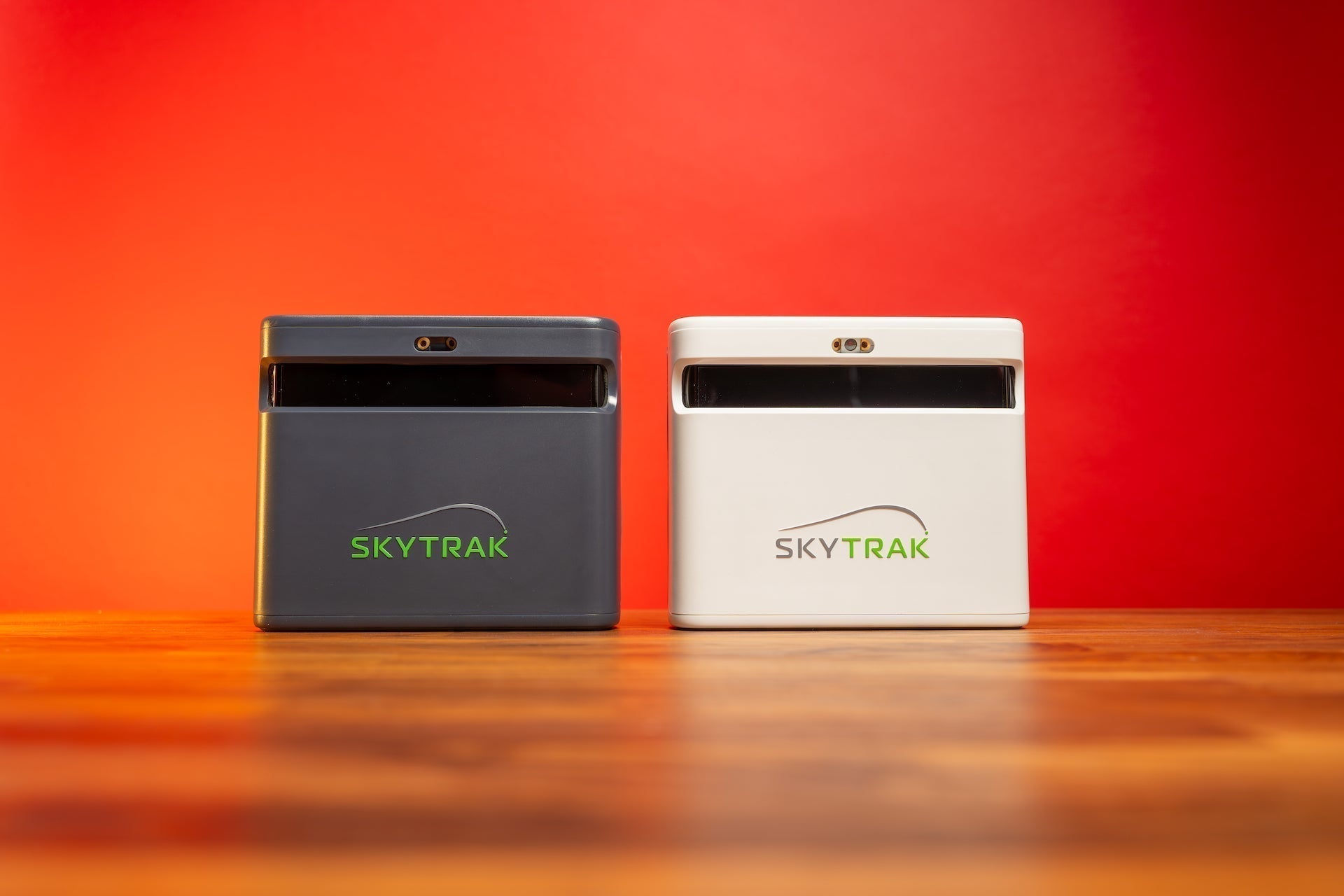 The gray ST MAX and white SkyTrak+ golf launch monitors side by side on a woodgrain surface against a red background