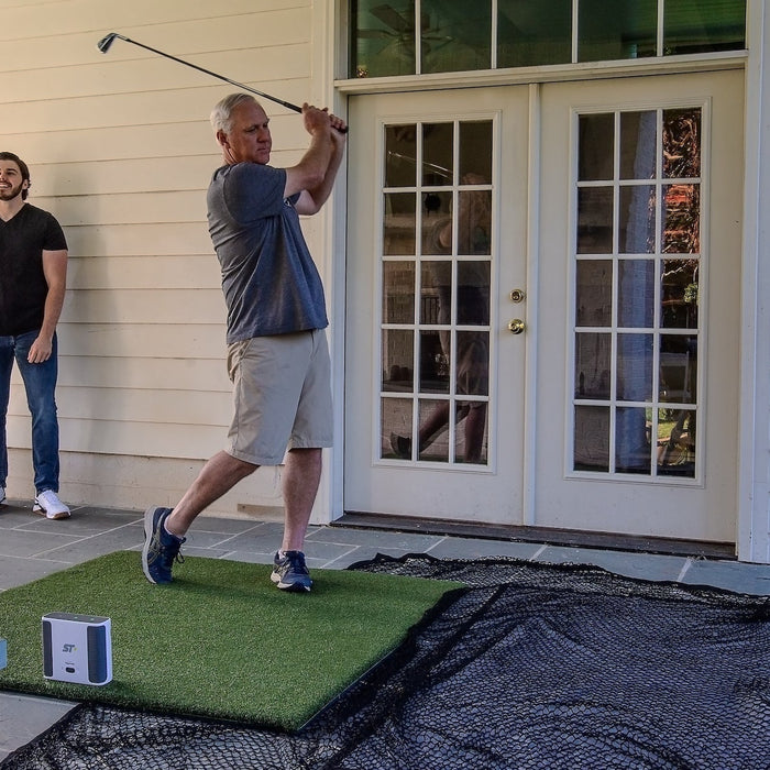 A senior golfer post swing in a SkyTrak+ home golf sim setup with a golf hitting mat, golf net and a man watching him