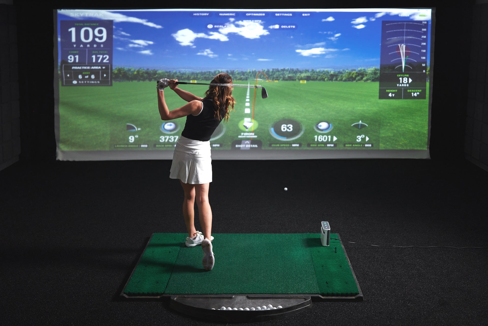 A woman post-swing on a golf hitting mat in a darkened golf simulator with the SkyTrak+ and SkyTrak software on the golf impact screen