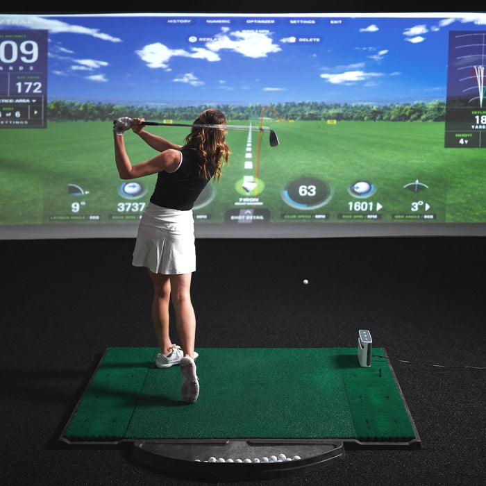 A woman post-swing on a golf hitting mat in a darkened golf simulator with the SkyTrak+ and SkyTrak software on the golf impact screen