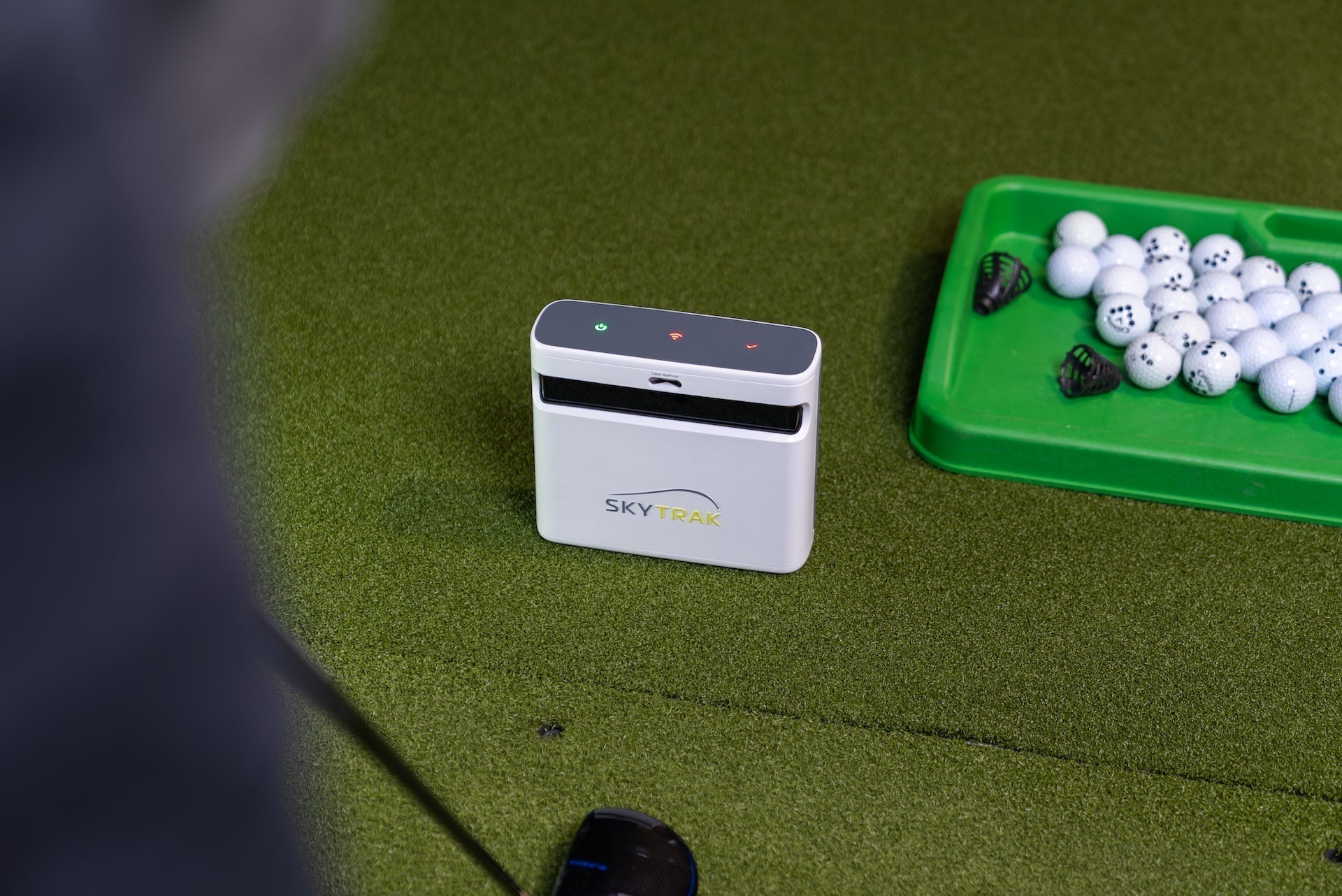A SkyTrak+ golf launch monitor on a golf hitting mat next to a tray of golf balls