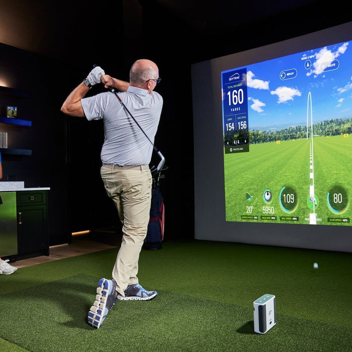 A man post-swing in a home golf simulator with a SkyTrak Plus launch monitor with SkyTrak software on the impact screen and a young lady leaning against a counter to the side