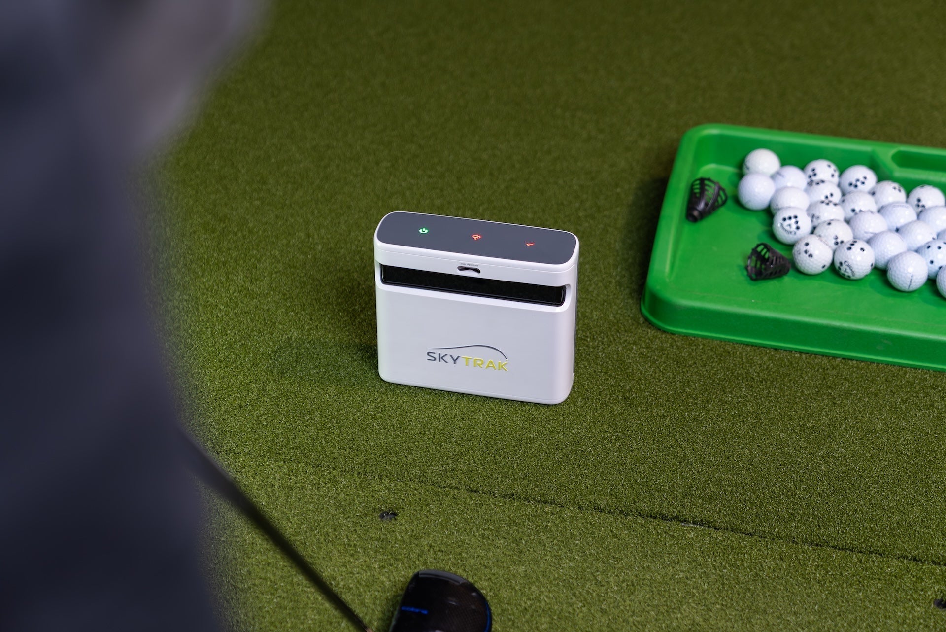 The SkyTrak+ viewed over the shoulder of a golfer in a golf simulator with a driver on a golf hitting mat next to a tray full of golf balls
