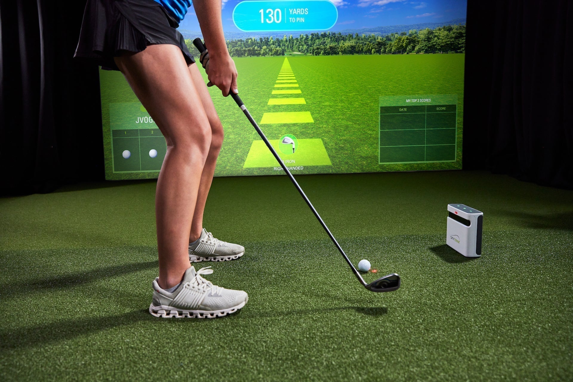 The lower half of a female golfer getting ready to hit a golf ball with an iron in front of a SkyTrak+ launch monitor in a home golf simulator