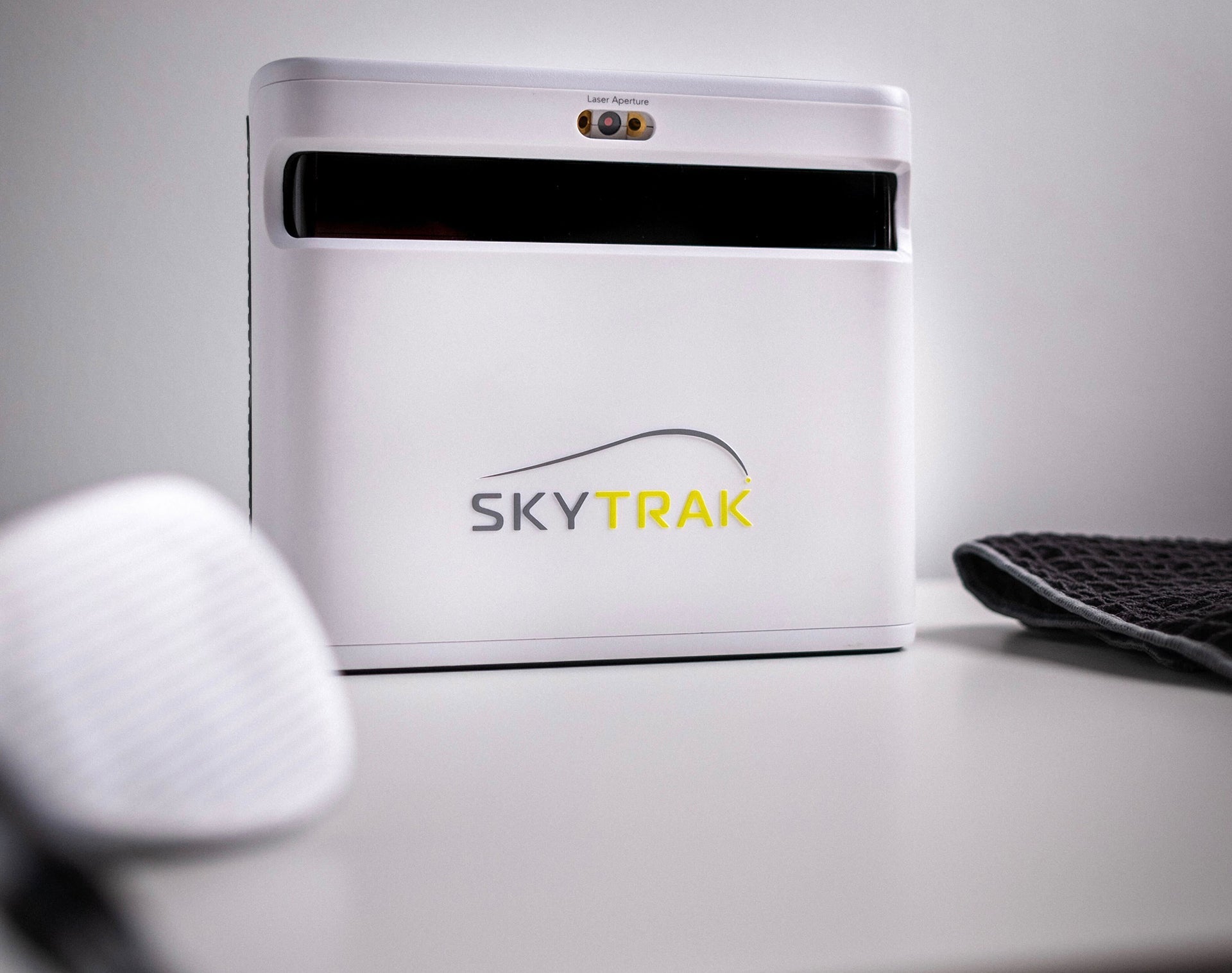 The SkyTrak+ launch monitor on a white table with a golf club head and golf towel on either side