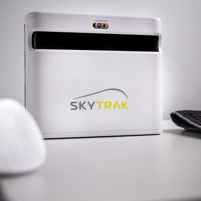 The SkyTrak+ launch monitor on a white table with a golf club head and golf towel on either side
