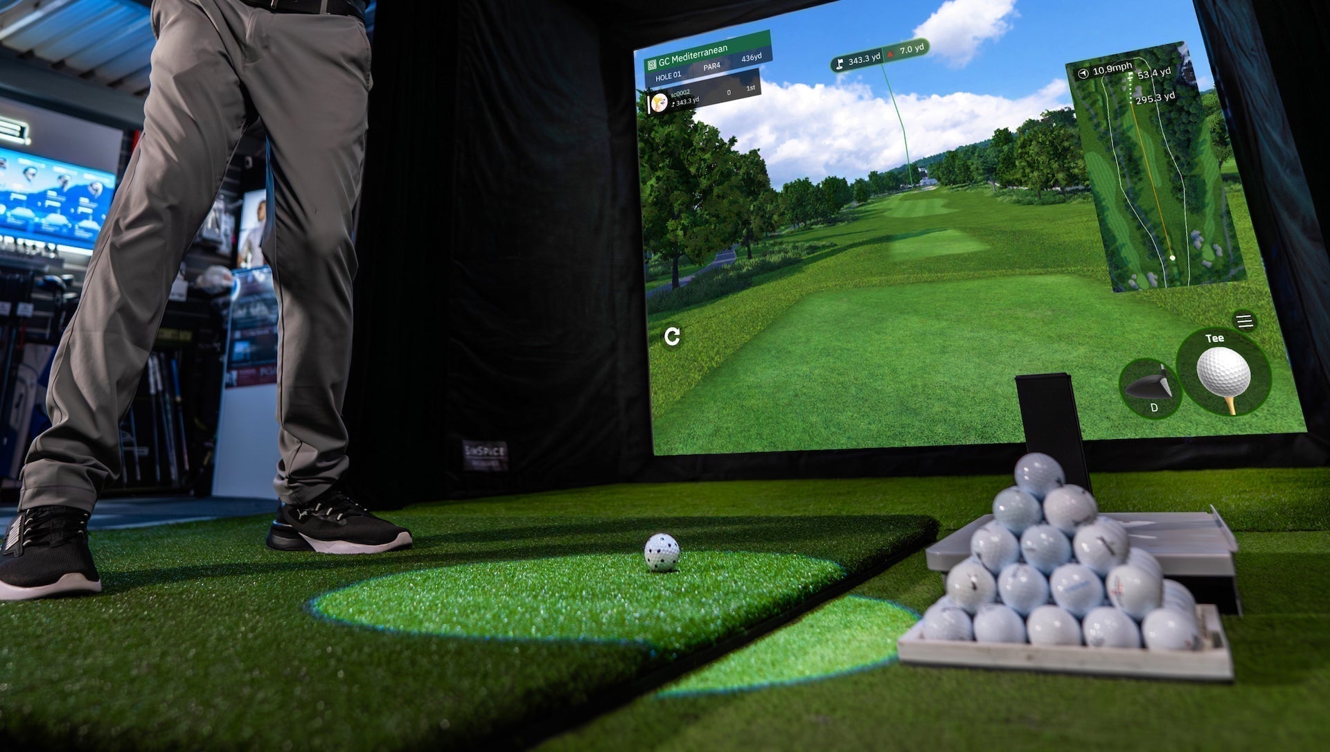 The lower half of a golfer in a golf simulator with a Square Golf launch monitor, a pyramid of golf balls on a golf hitting mat and a golf impact screen with golf course simulation on it