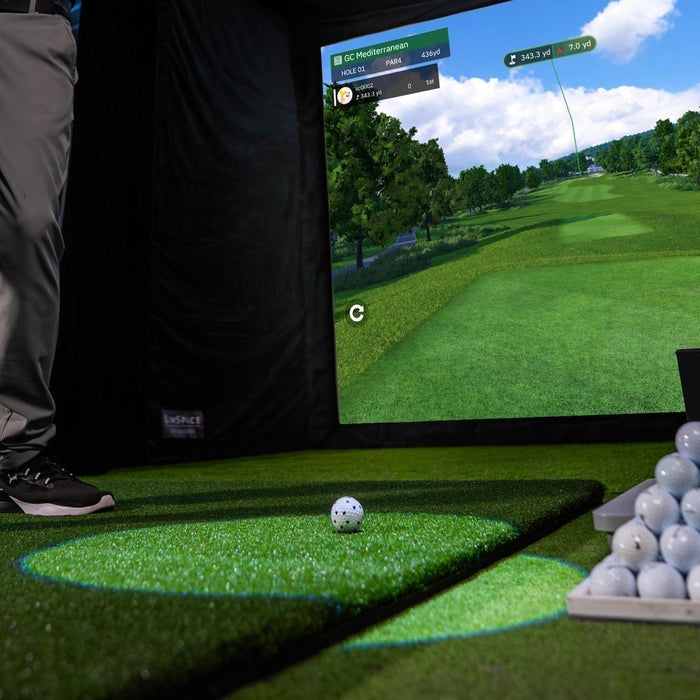 The lower half of a golfer in a golf simulator with a Square Golf launch monitor, a pyramid of golf balls on a golf hitting mat and a golf impact screen with golf course simulation on it