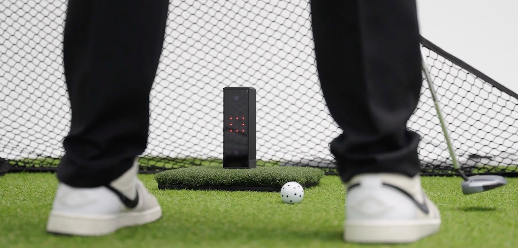 The Square Golf Launch Monitor between the feet of a golfer with a putter to the right and a golf net behind the unit