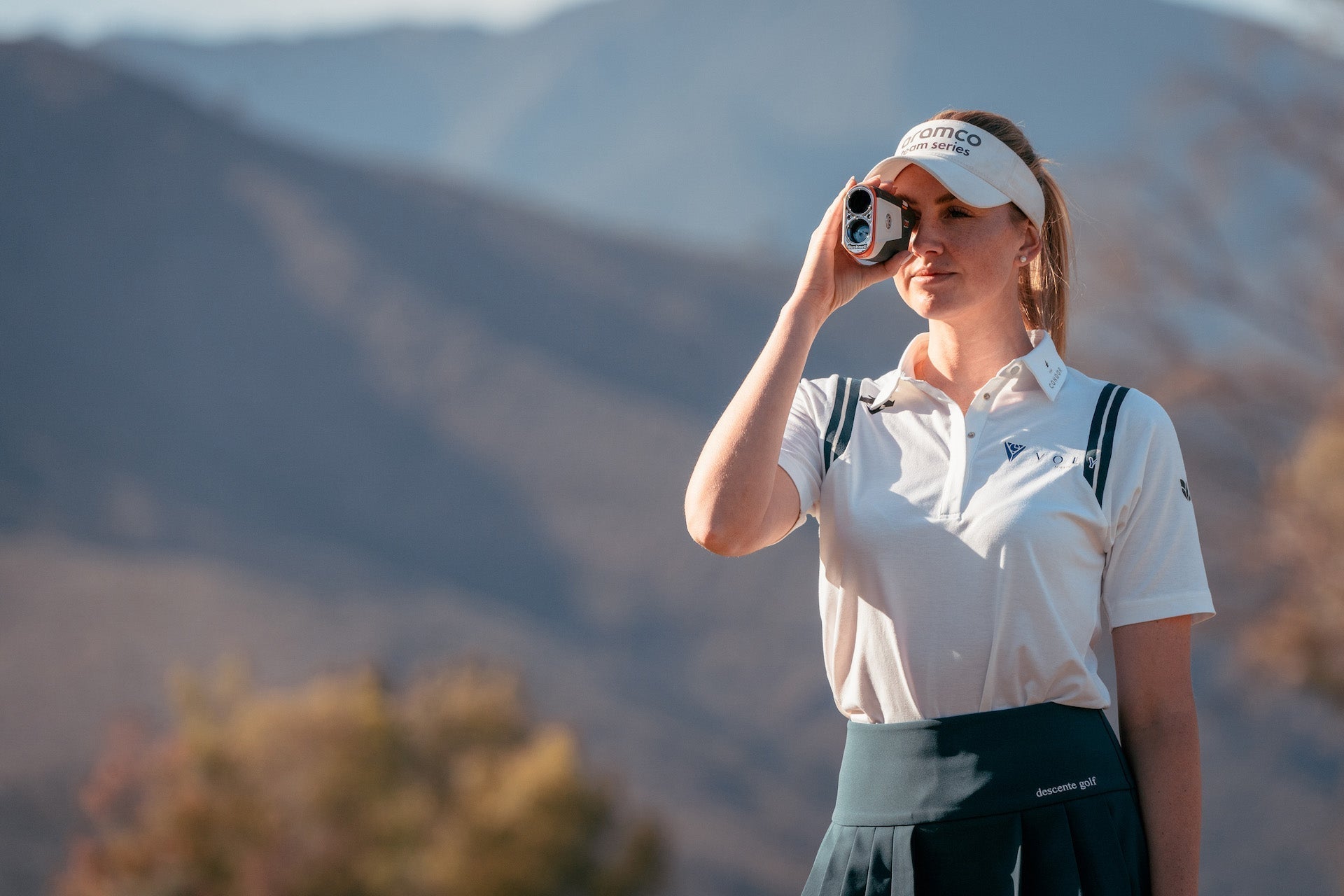 A woman in golf wear holding a Bushnell rangefinder to her eye on a golf course with a mountain range in the background