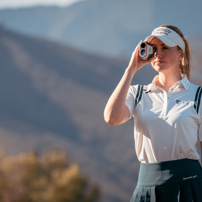 A woman in golf wear holding a Bushnell rangefinder to her eye on a golf course with a mountain range in the background