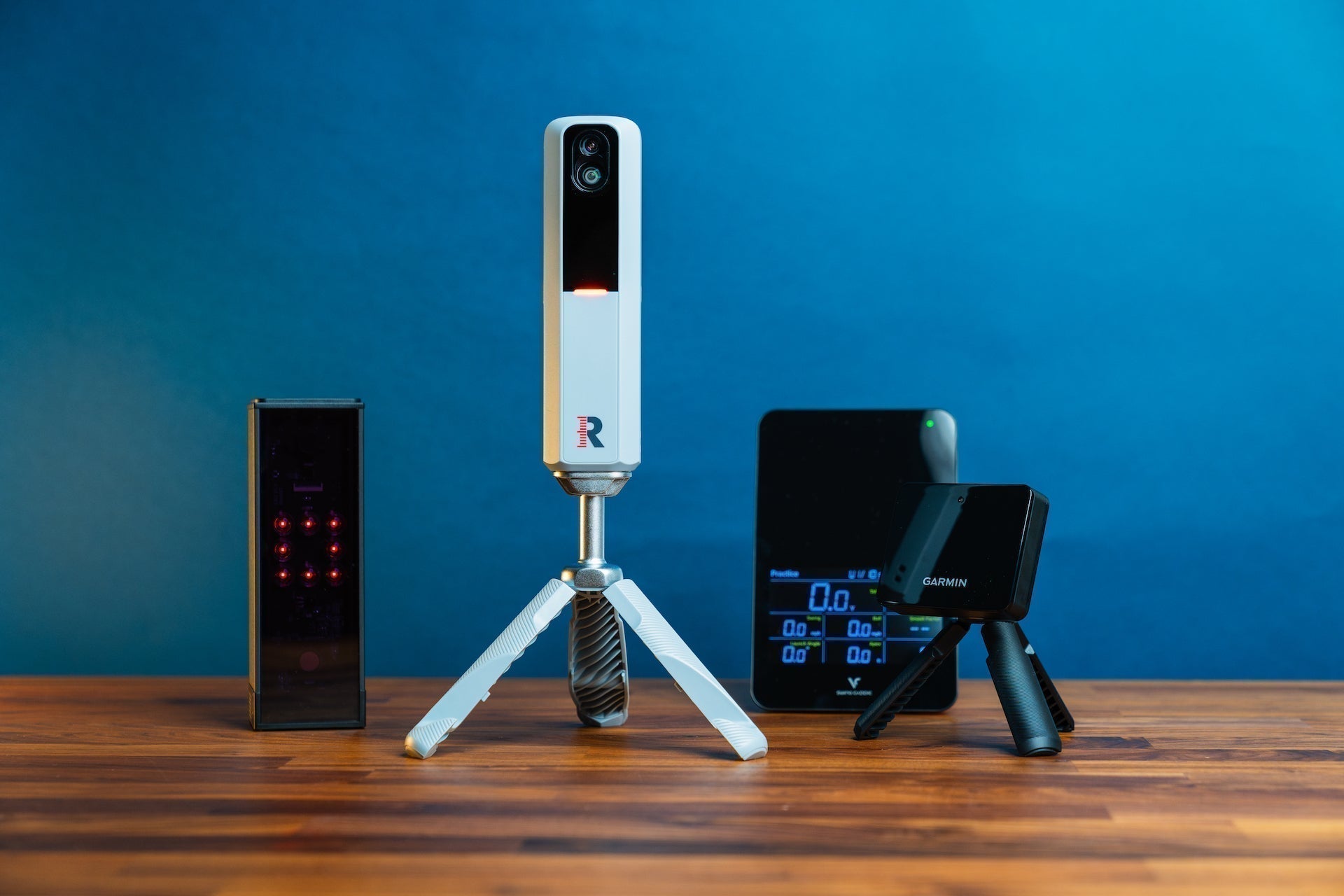 Group photo of Square, Rapsodo MLM2PRO, Swing Caddie SC4 Pro and Garmin R10 on a woodgrain surface against a blue background