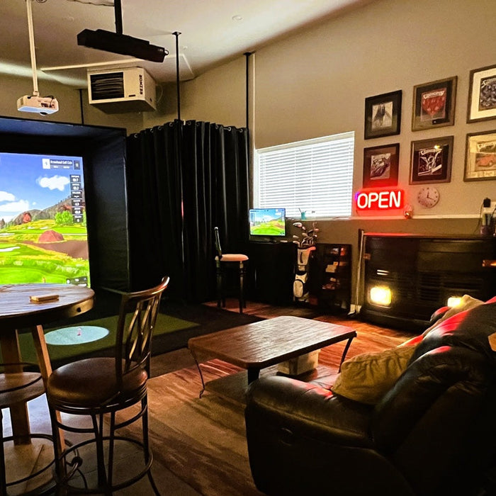 A home golf simulator in a entertainment room with the Uneekor Eye XO overhead golf launch monitor, golf hitting mat, golf impact screen and enclosure with a projector hanging from the ceiling with simulated golf course on the screen