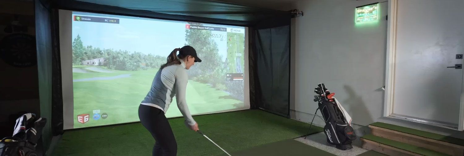 A woman getting ready to swing in a garage golf simulator with a SportScreen Vanish retractable enclosure