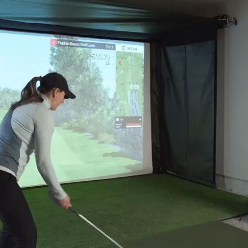 A woman getting ready to swing in a garage golf simulator with a SportScreen Vanish retractable enclosure