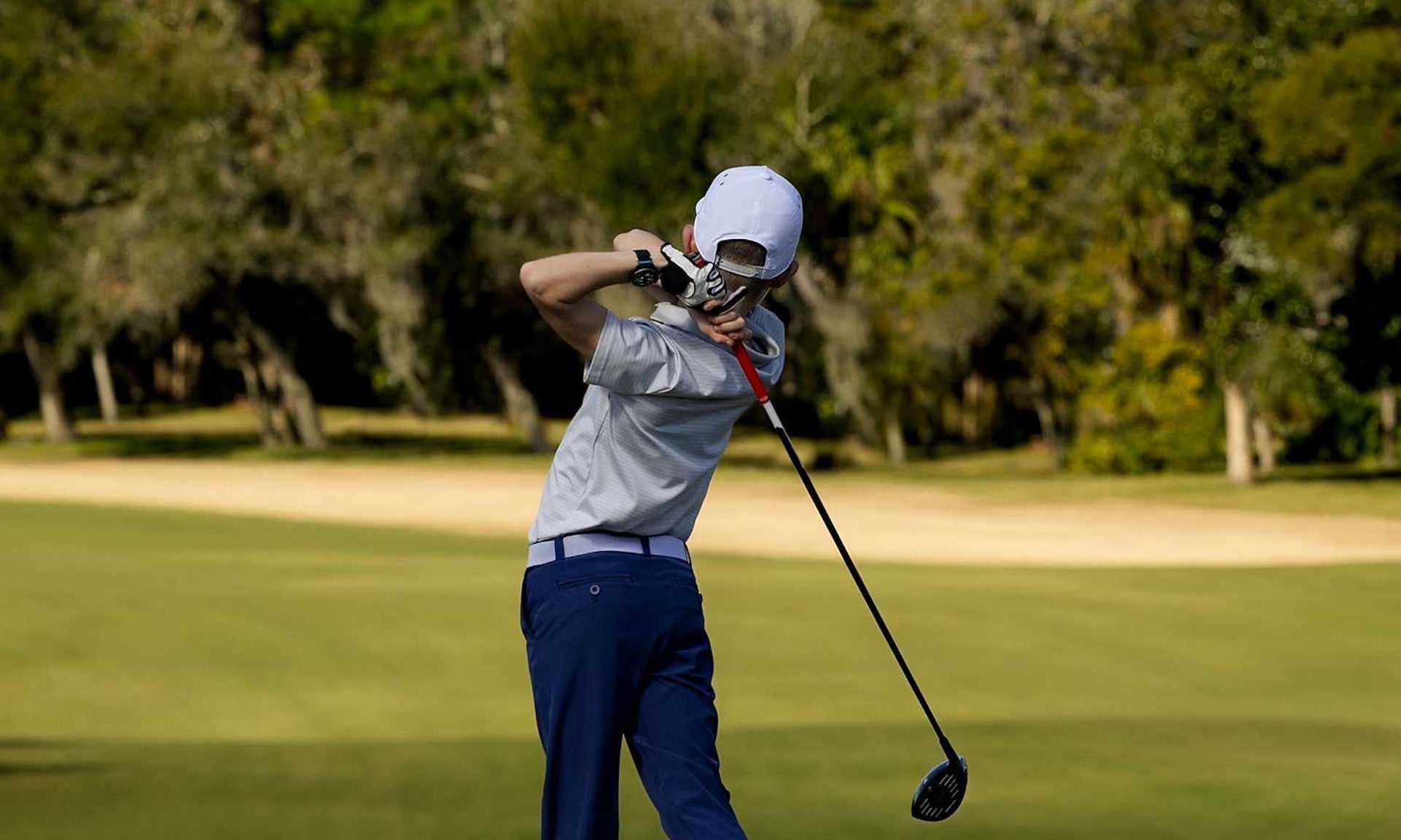 A young golfer post swing at the tee on a golf course wearing the Garmin Approach J1 golf GPS watch