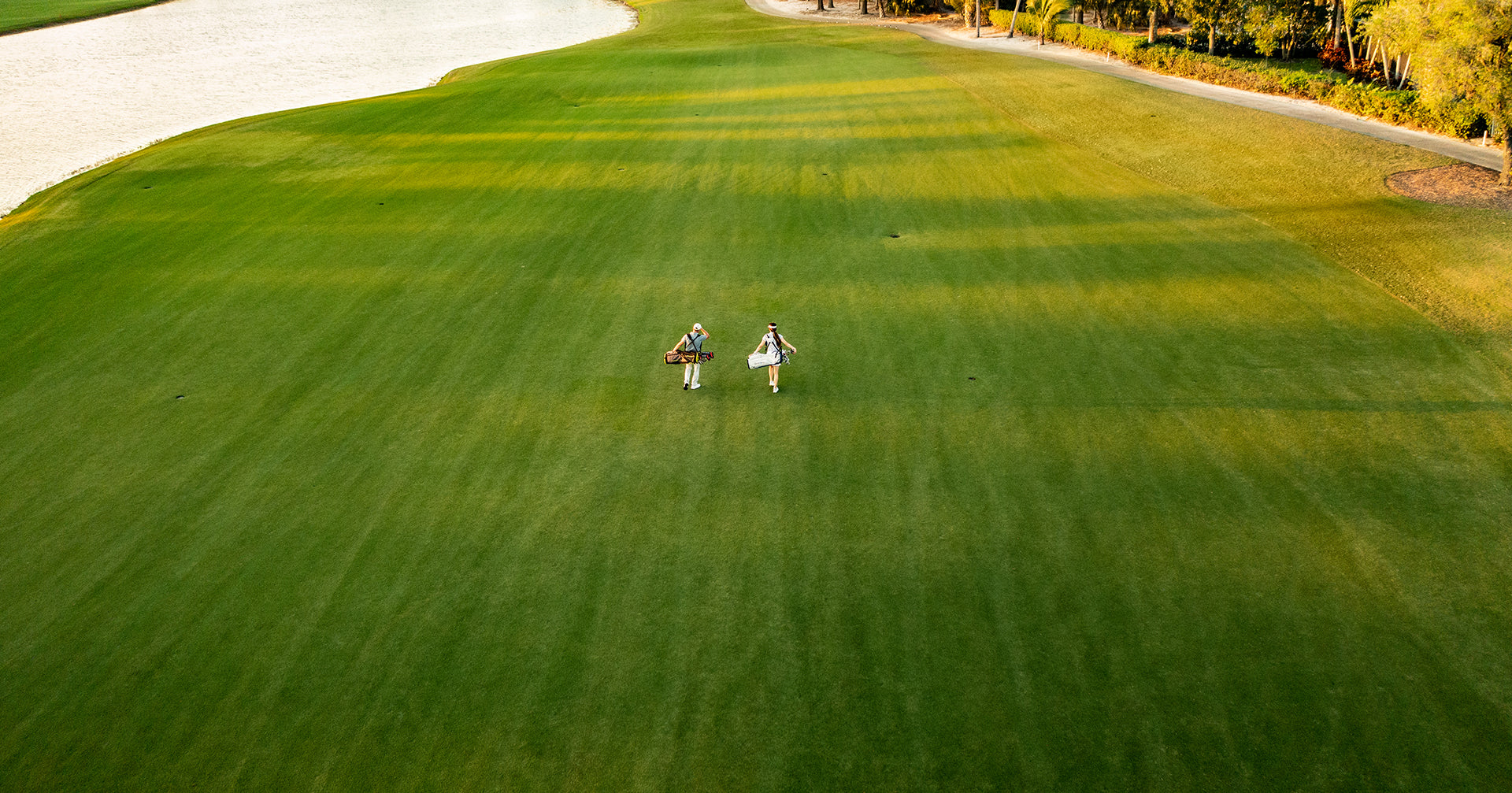 A wide shot of a golf fairway with two people walking up the middle and a sand trap on the left