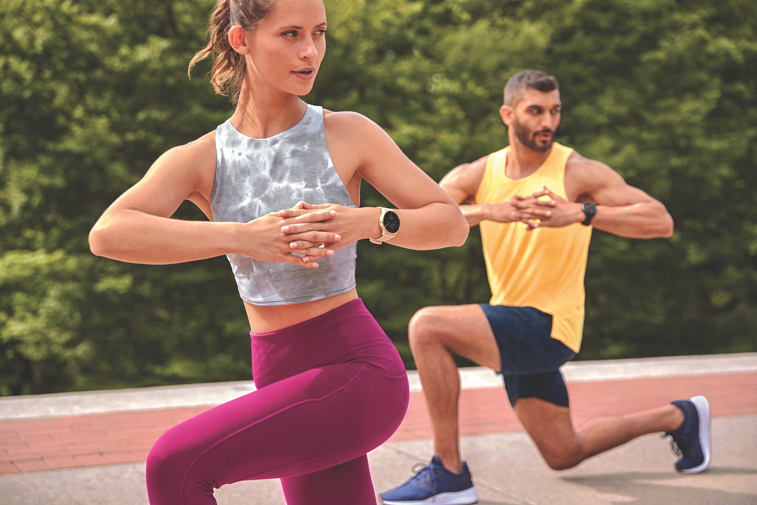 A woman and man doing knee bends outdoors while wearing Garmin Venu 2 Plus watches